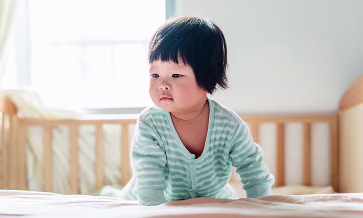 Baby in striped onesie crawling on a bed in a bright room.