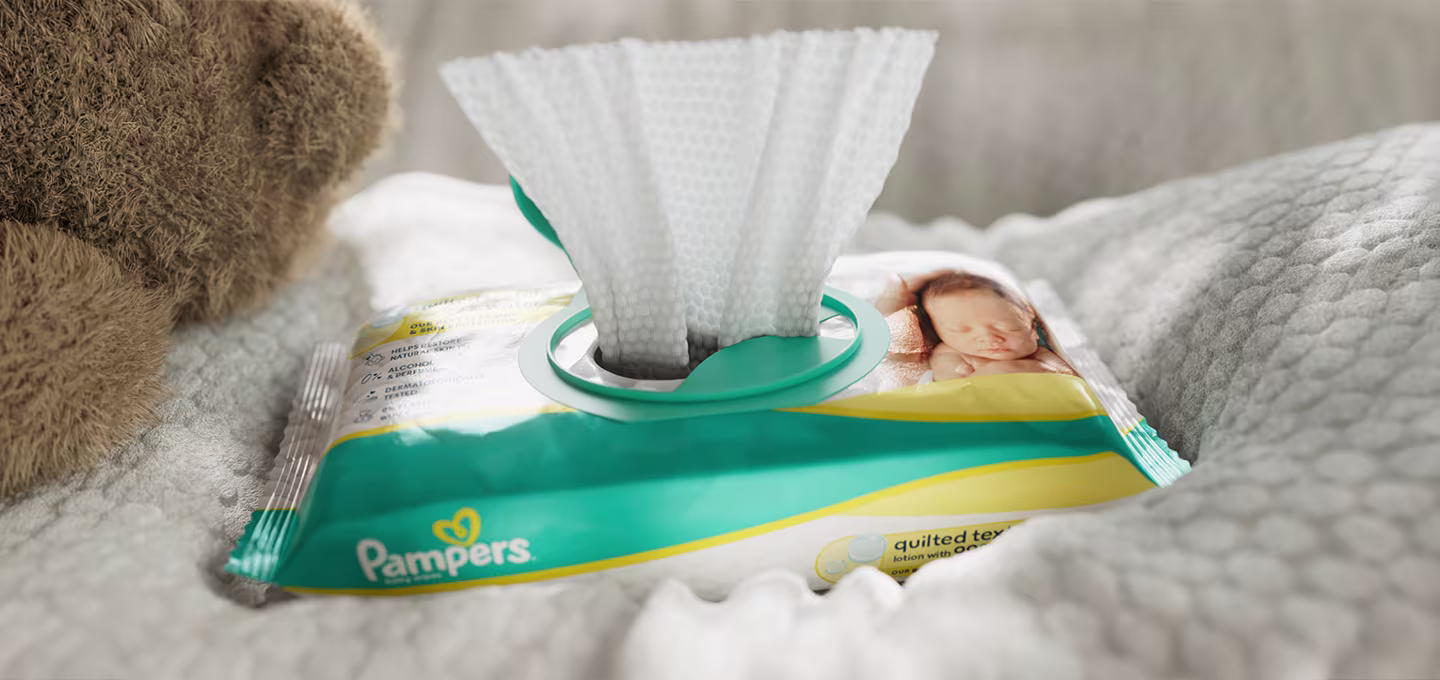 Pampers baby wipes packet with a wipe pulled through the lid, resting on a soft blanket beside a teddy bear