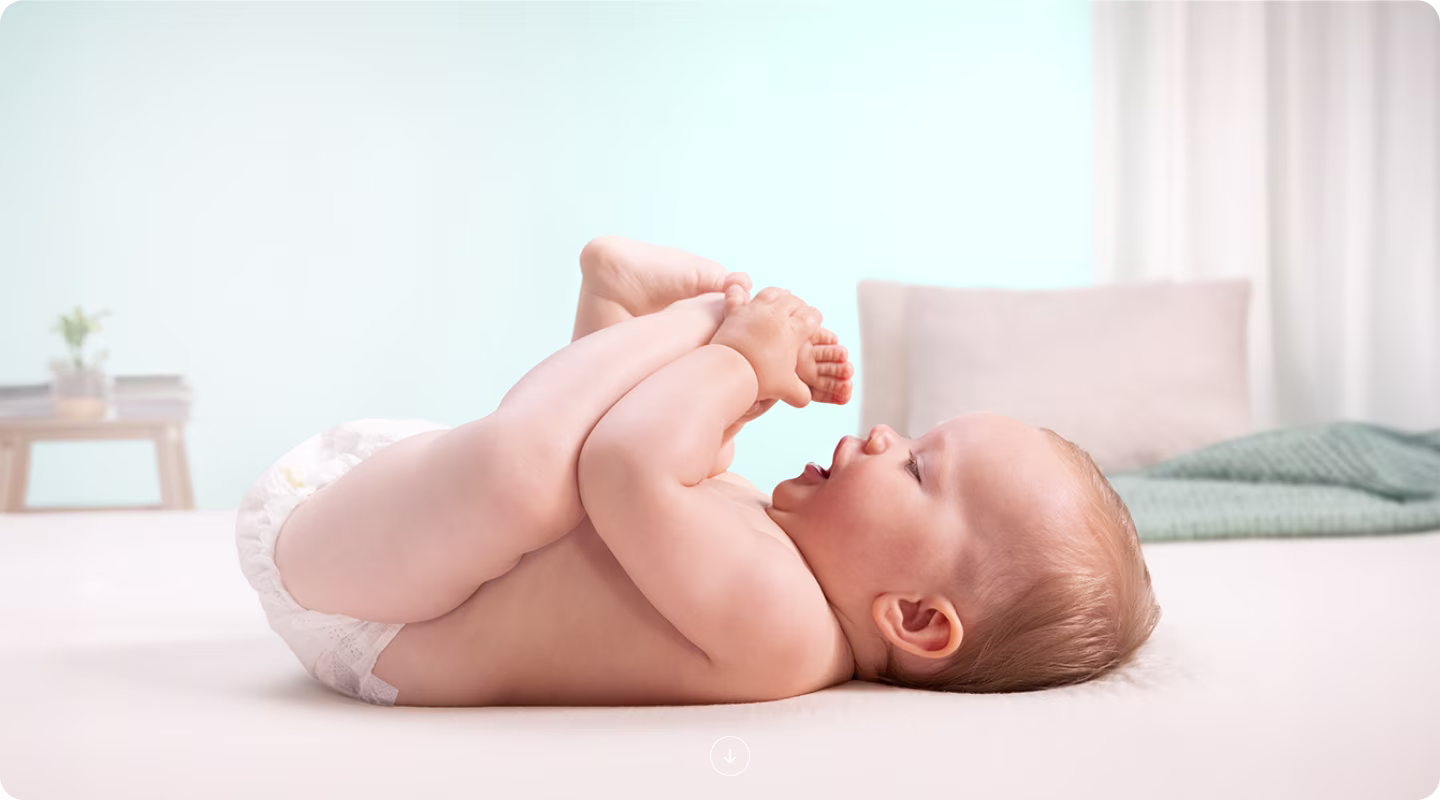 A baby lies on their back on a white surface, holding its feet up.
