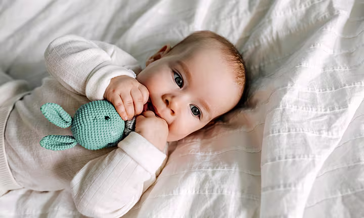 Baby liegt auf einem weichen Bett und hält ein gestricktes Spielzeug, auf dem es kaut.