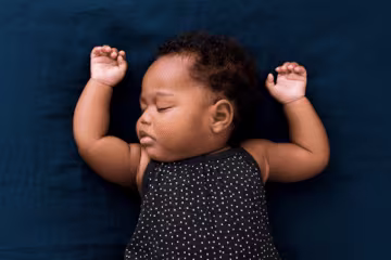 Sleeping baby in polka dot outfit on blue blanket, arms raised peacefully.