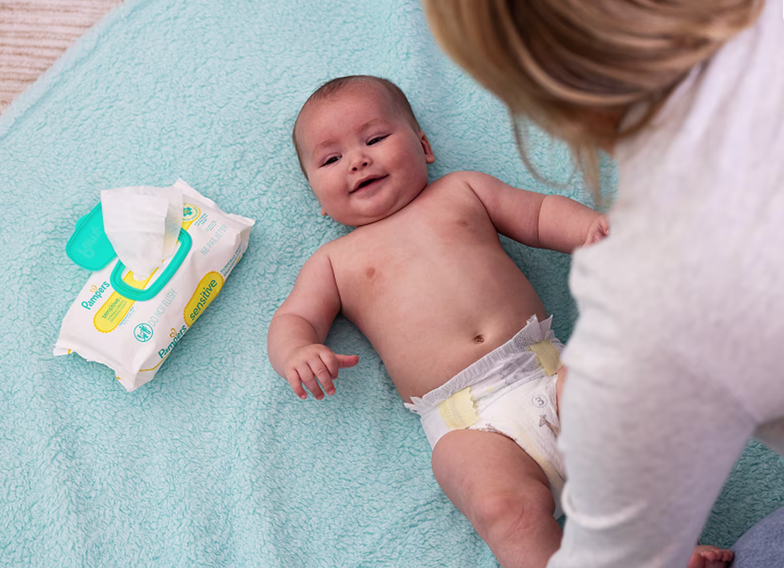 Smiling baby lays on their back on a soft blue blanket with a pack of sensitive wipes laying next to them and mom looking down on them