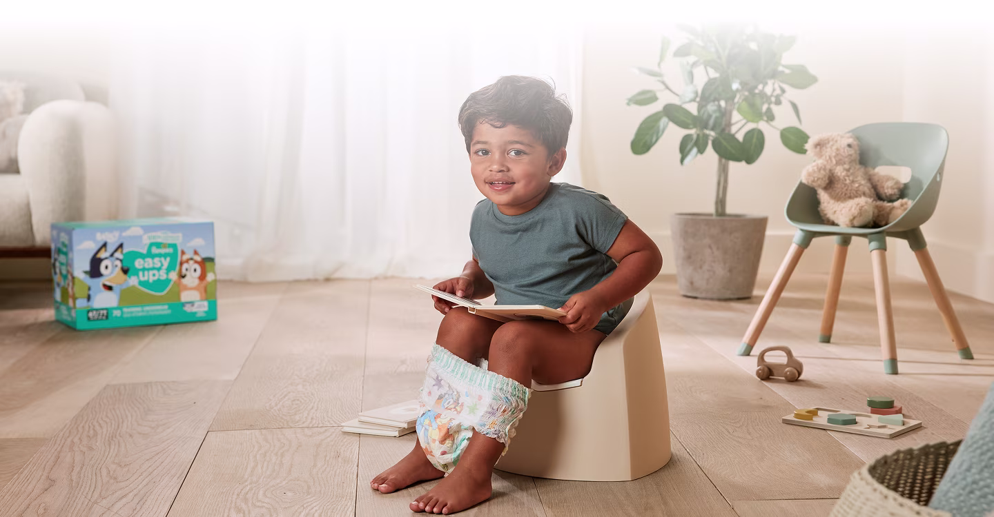 A toddler sits on a small, colorful potty chair in a brightly lit bathroom.