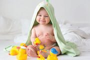 Smiling baby wrapped in a green towel playing with rubber ducks on a bed.