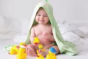 Smiling baby wrapped in a green towel playing with rubber ducks on a bed.