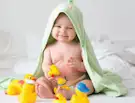 Smiling baby wrapped in a green towel playing with rubber ducks on a bed.