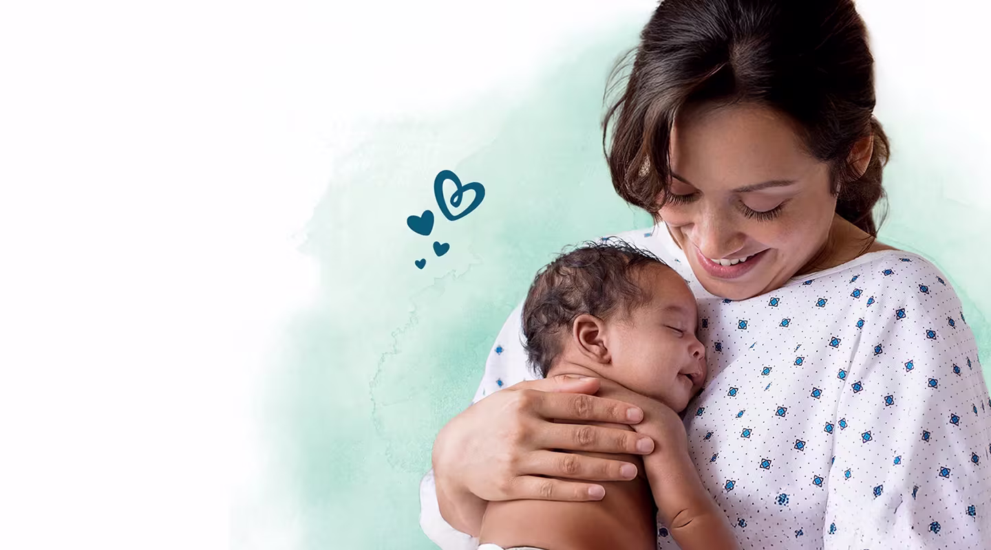 Una madre sonriente vestida con una bata de hospital sostiene a un bebé recién nacido dormido cerca de su pecho.