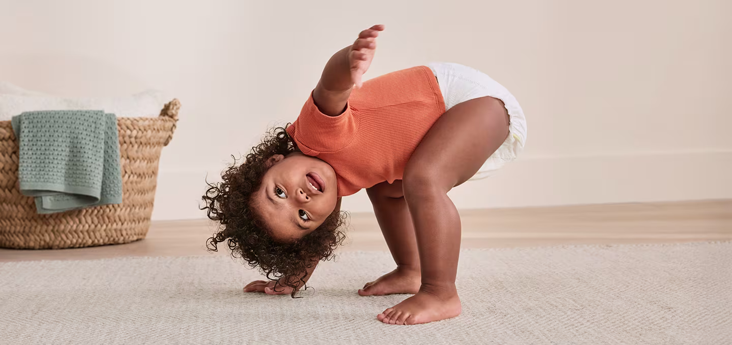 Toddler in a diaper and orange shirt bends playfully on a rug in a bright living room.