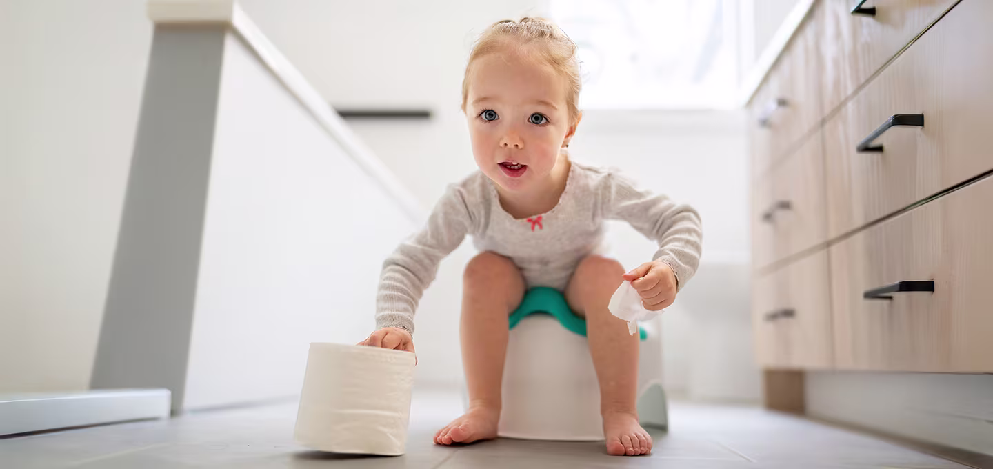 A toddler sits on a small training potty in a bright bathroom, holding toilet paper and looking up toward the camera.