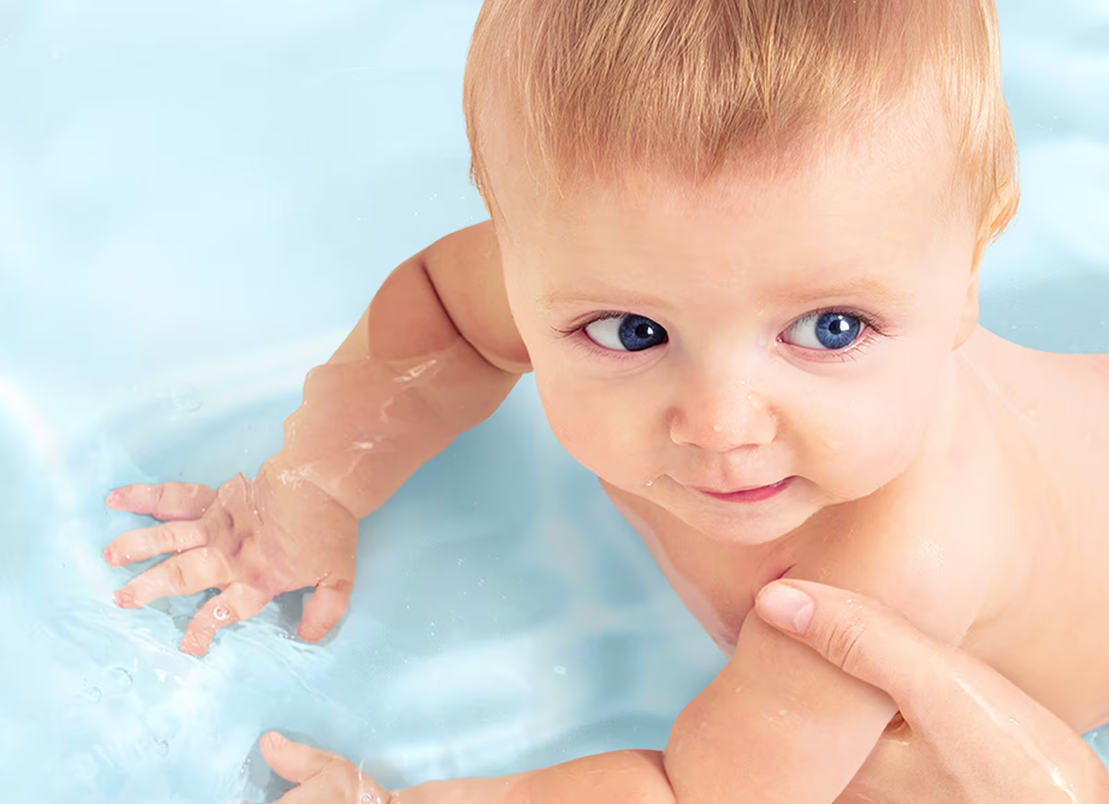 Eine Draufsicht auf ein Baby, das sanft in klarem, blauem Wasser von einer Hand eines Erwachsenen gestützt wird.