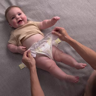 A smiling baby lying on their back during a diaper change. A parents hand is stretching the diaper sides to fasten the new diaper. The baby is dressed in a yellow t-shirt. All sensitive parts are covered.