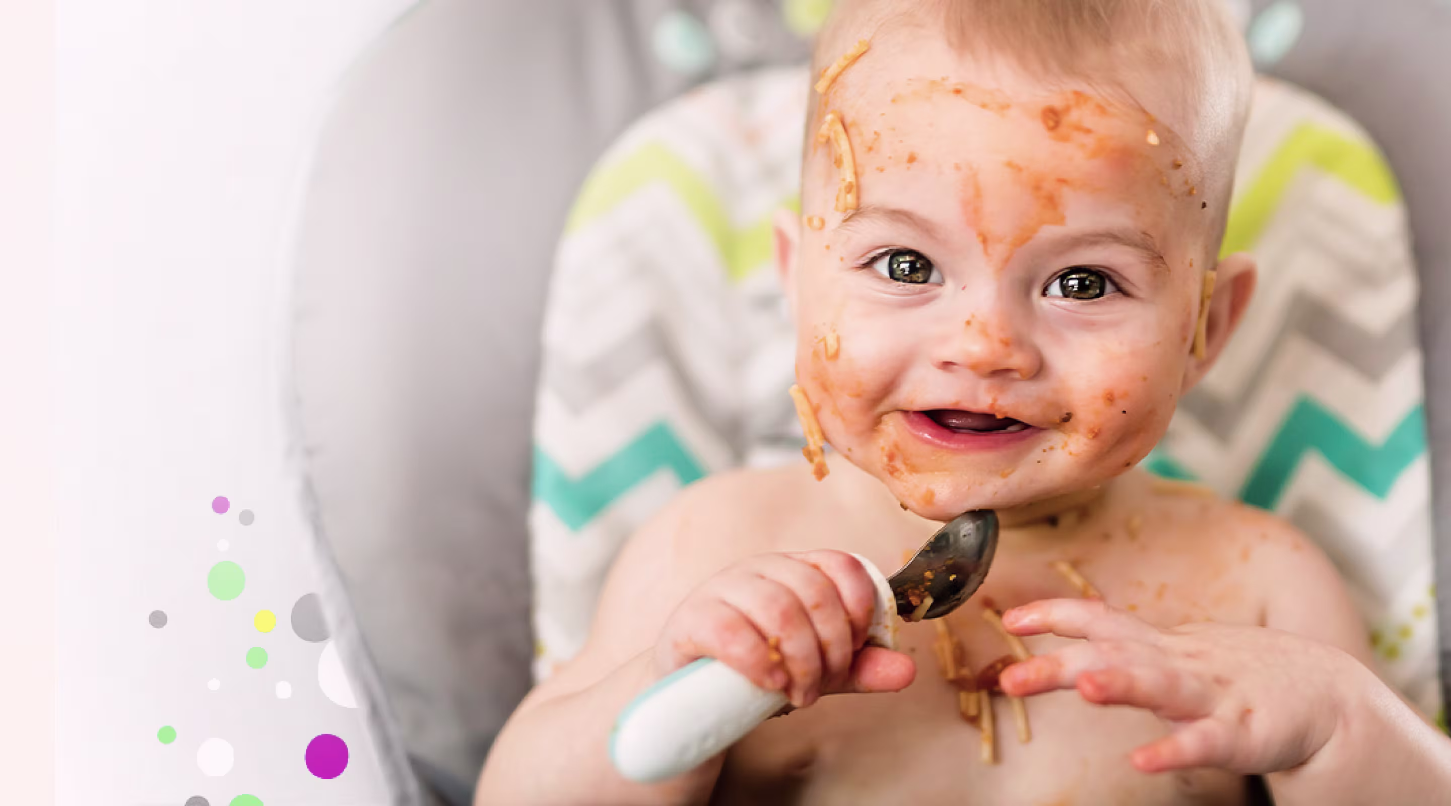 Lächelndes Baby im Hochstuhl, Gesicht mit Spaghetti-Sauce bedeckt, hält einen Löffel