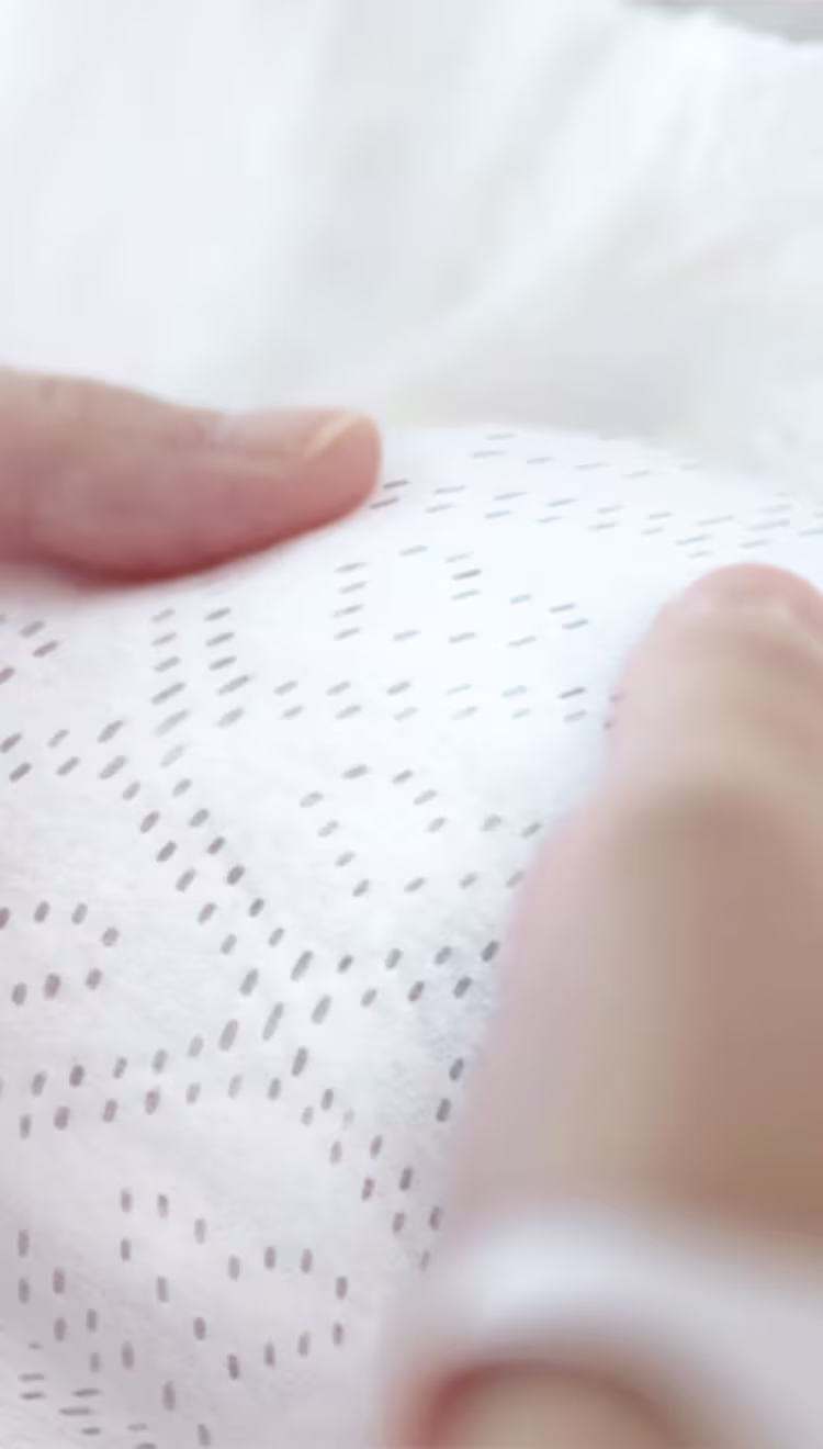 Eine Nahaufnahme zeigt zwei Hände, die sanft das Pampers Windelinnenvlies halten, wobei die Struktur und die Weichheit verdeutlicht werden.