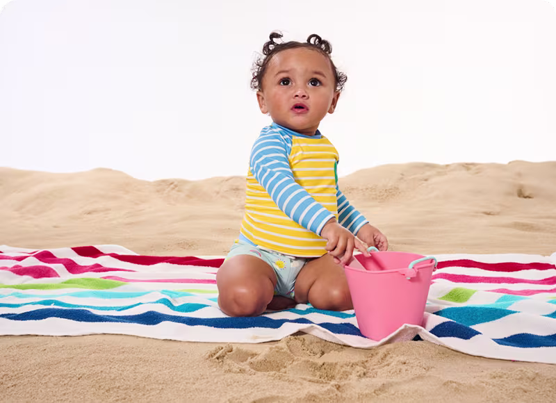 A baby sitting on a towel in the sand with a bucket, enjoying a sunny day at the beach