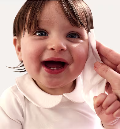 Um primeiro plano do rosto de um bebé enquanto uma mão lhe limpa suavemente a bochecha com uma toalhita Dodot Sensitive. Um primeiro plano do rosto de um bebé enquanto uma mão lhe limpa suavemente a bochecha com uma toalhita Dodot Sensitive.