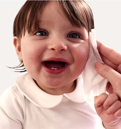 Um primeiro plano do rosto de um bebé enquanto uma mão lhe limpa suavemente a bochecha com uma toalhita Dodot Sensitive. Um primeiro plano do rosto de um bebé enquanto uma mão lhe limpa suavemente a bochecha com uma toalhita Dodot Sensitive.