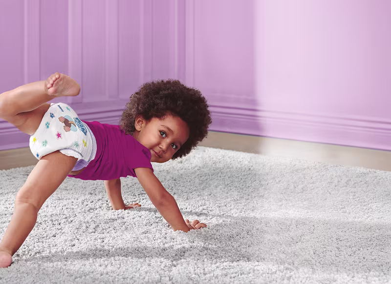 Um bebé num chão alcatifado numa sala roxa, vestido com uma fralda branca com desenhos coloridos de animais de desenhos animados.