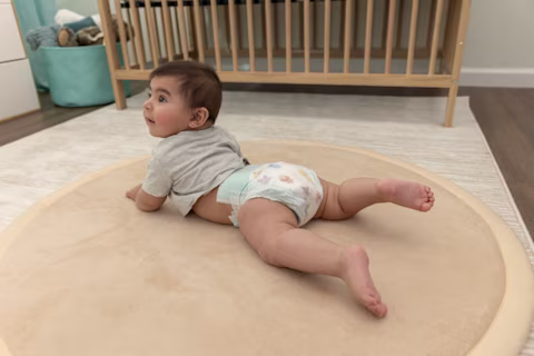 Un bebé está acostado boca abajo sobre una alfombra en su habitación, mientras lleva puesta una camiseta gris y un pañal Pampers Baby Dry.