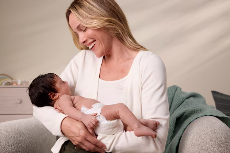 Une mère assise tient son nourrisson en couche dans ses bras et lui sourit affectueusement.