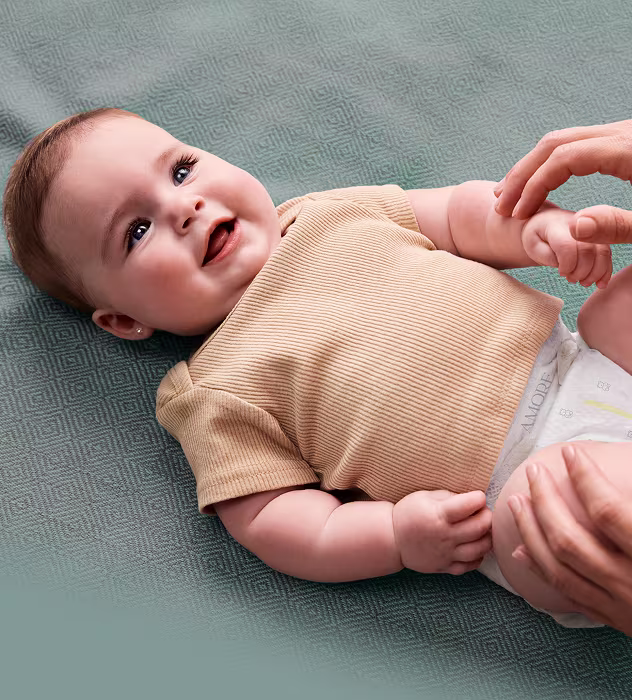 Baby laying on a teal sheet smiling up at parent while wearing an amore diaper and cream shirt