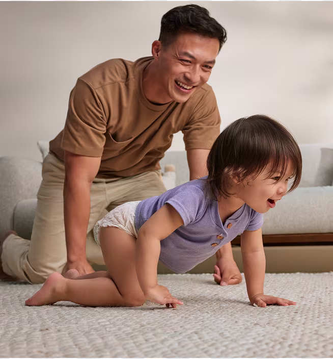 Father and baby crawling after each other on a grey carpet smiling. Baby is wearing a violet tshirt and pampers diapers. father dressed in beige tones