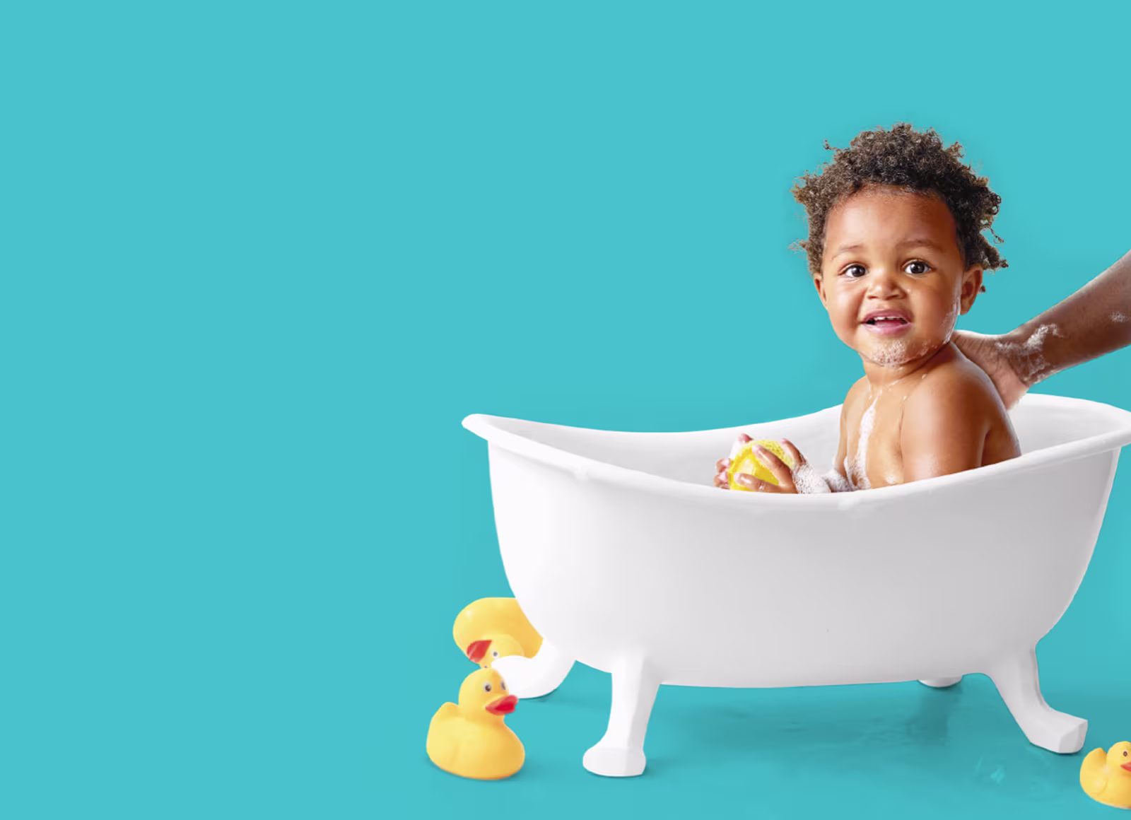 Ein glückliches Kleinkind sitzt in einer kleinen weißen Badewanne. Eine Hand eines Erwachsenen wäscht sanft den Rücken des Kleinkindes, während gelbe Gummienten auf dem Boden daneben sitzen.