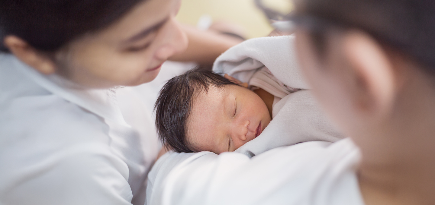 A newborn wrapped in a blanket rests calmly in a caregiver’s arms, showing healthy skin tone as adults gently observe for signs of jaundice.