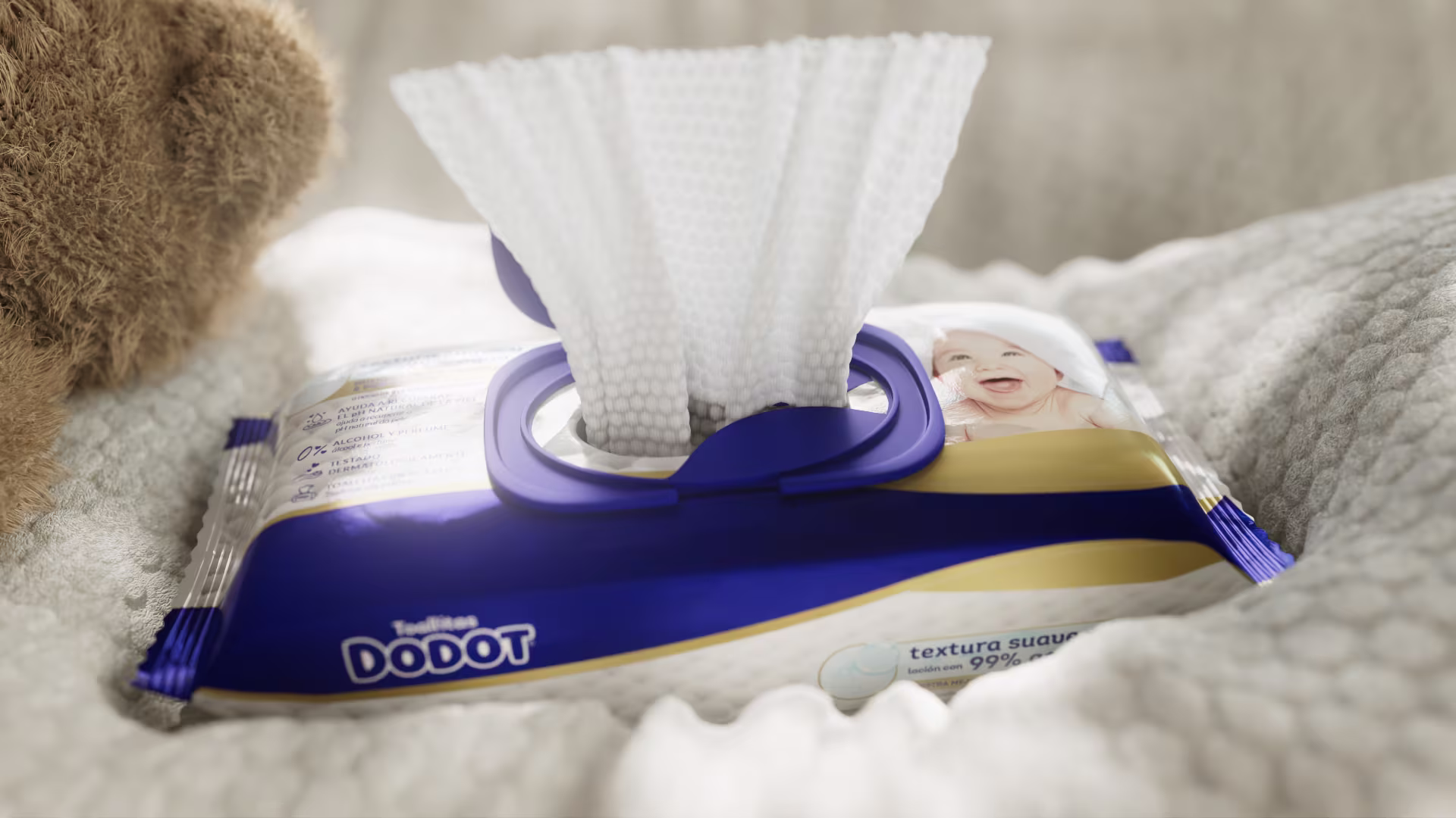 Toallitas húmedas Dodot con una toallita extraída del envase, sobre una manta suave junto a un oso de peluche.