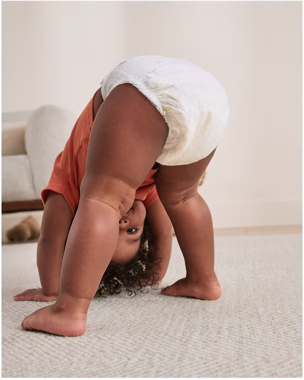 A baby playing in the living room doing a wild child pose on all fours whilst dressed in an orange shirt and pampers diaper pants.