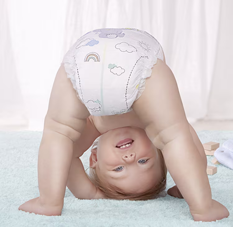 Ein Baby in einer bedruckten Windel beugt sich spielerisch über einen hellblauen Teppich und lächelt kopfüber. Die Szene ist hell und fröhlich und strahlt Freude und Unschuld aus.