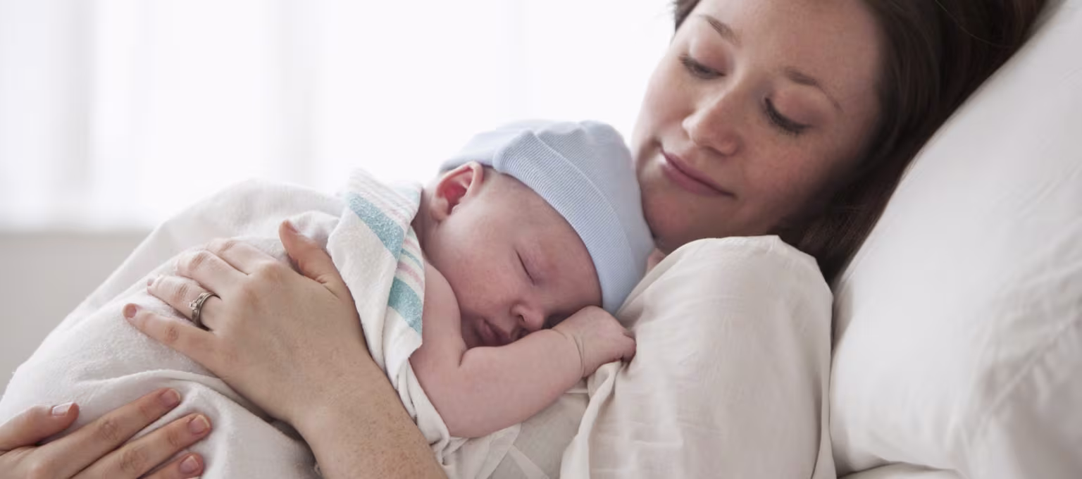 Madre recostada sosteniendo a su recién nacido dormido sobre el pecho, ilustrando posiciones de lactancia cómodas y contacto piel con piel.