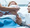 A mother in a hospital bed receives her newborn from a healthcare professional, with her partner beside her.