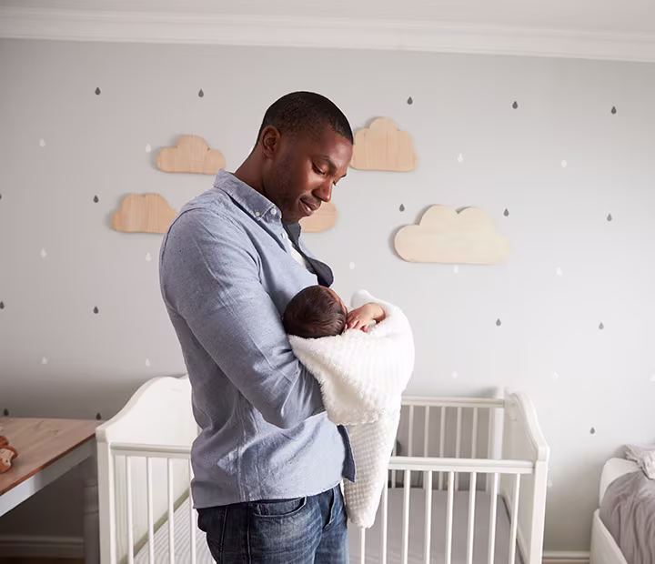 Father gently holding a newborn wrapped in a blanket, standing in a cozy nursery with cloud decorations.