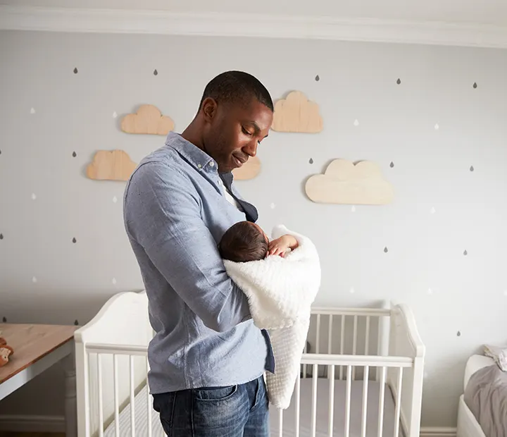 Nursery Vibes! What’s Your Dream Baby Room Style? Father gently holding a newborn wrapped in a blanket, standing in a cozy nursery with cloud decorations.