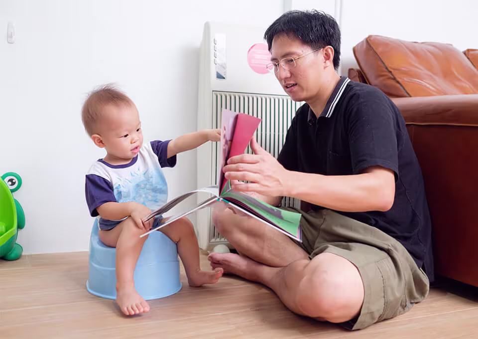 A toddler sitting on a potty while his father shows him a book