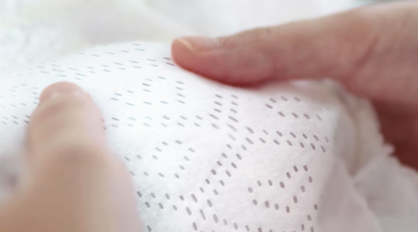 Et nærbilde viser to hender som forsiktig holder en myktexturert Pampers-bleie, noe som fremhever dens prikkete mønster og plysjfølelse.