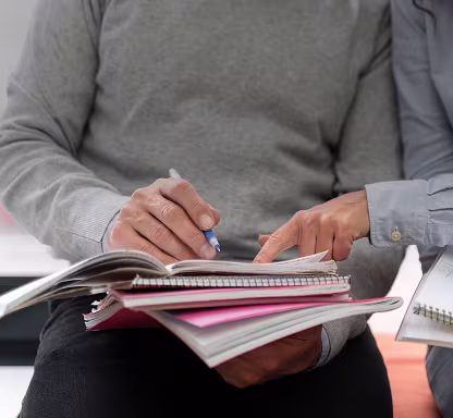 Two people sit close together, reviewing open notebooks and pointing at notes during a study session.