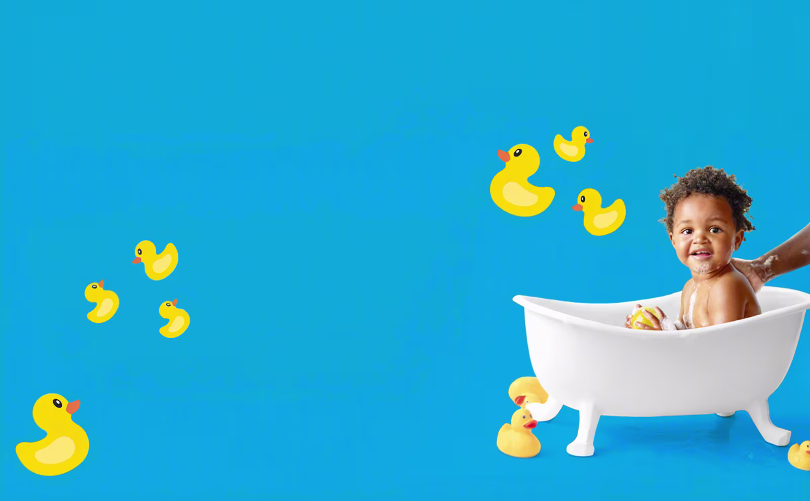 Un feliz niño pequeño se sienta en una bañera blanca en miniatura, sonriendo a la cámara con burbujas de jabón en la cara. La mano de un adulto lava suavemente la espalda del niño, mientras patitos de goma amarillos están en el suelo cerca.
