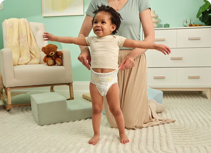 Un niño pequeño con una camiseta naranja de canalé camina en su sala de estar mientras lleva puesto un pañal Swaddlers 360° tipo pull on.