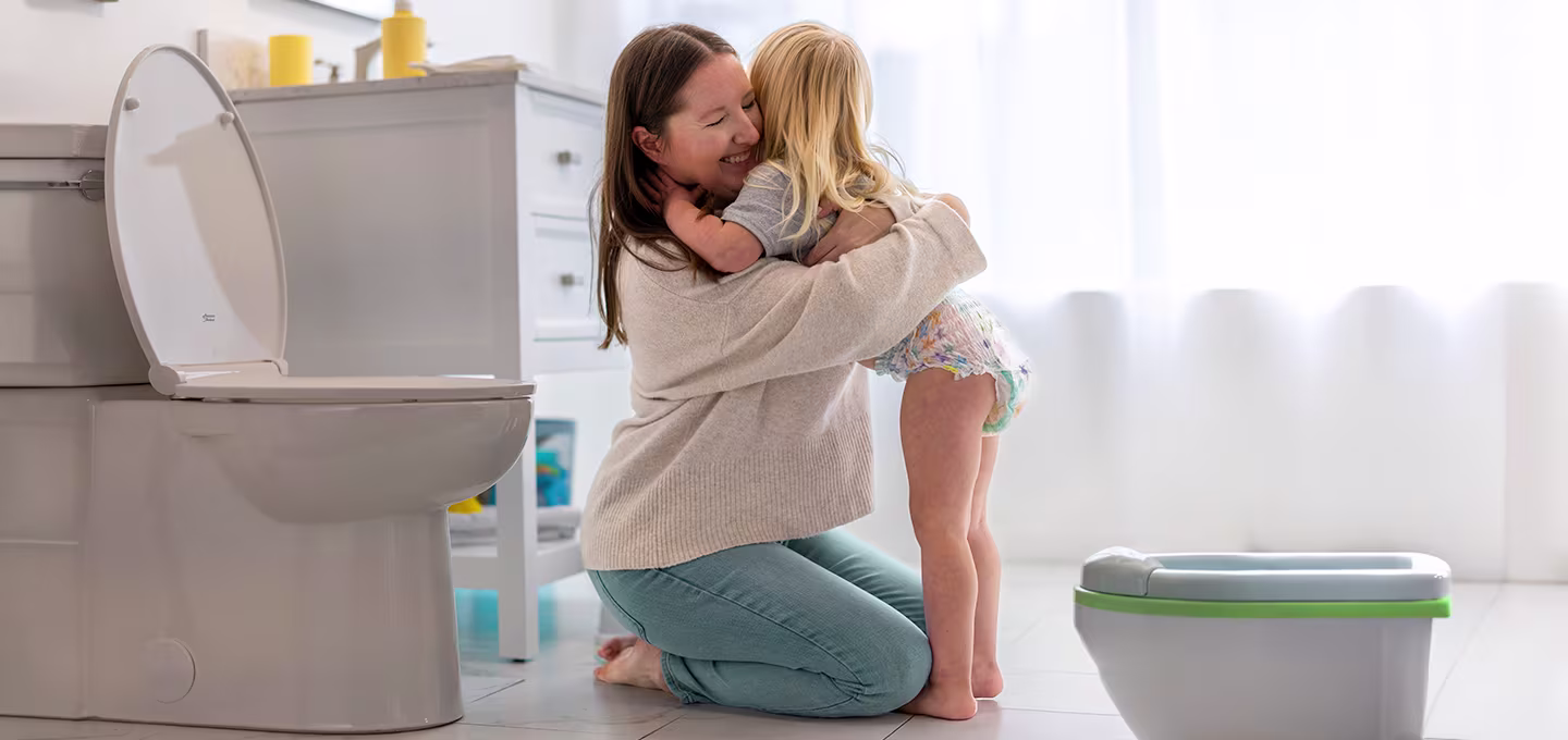 Mother kneeling in a bright bathroom hugs her toddler in a diaper beside a toilet and child potty.