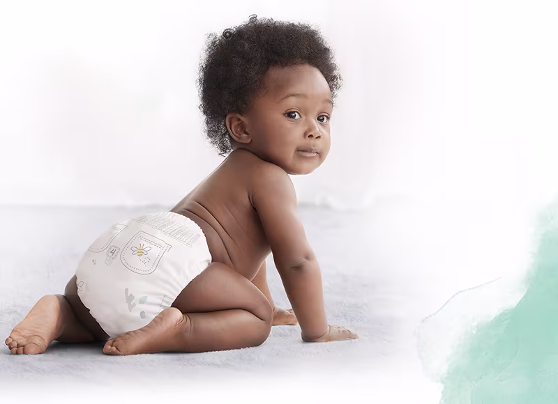 Suloinen vauva, pukeutuneena valkoiseen vaippaan jossa on mehiläis- ja taskukuvio, on kuvattu takaapäin kun hän ryömii pehmeällä matolla.