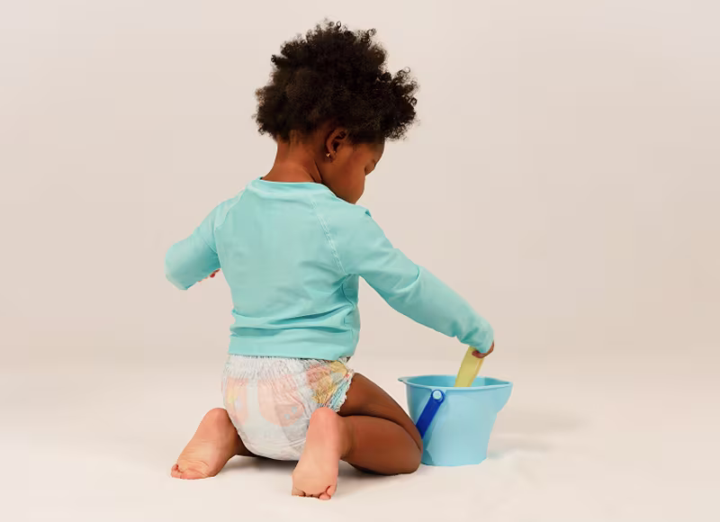 A toddler wearing Pampers Splashers and a teal swim shirt kneels down while playing with a spade and pail