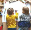 Two young children drawing on paper surrounded by colorful toys and pencils on a wooden floor.