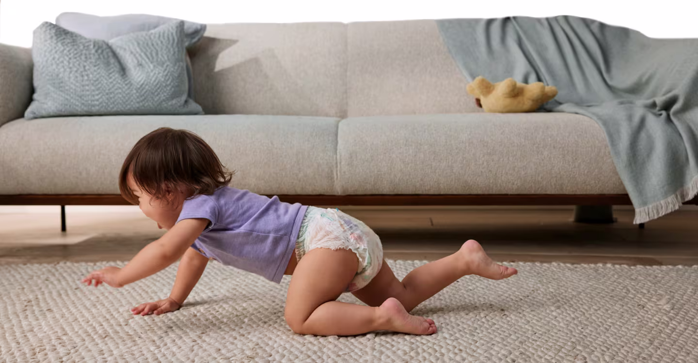 A baby crawls on a light-colored rug with a gray sofa visible in the background.