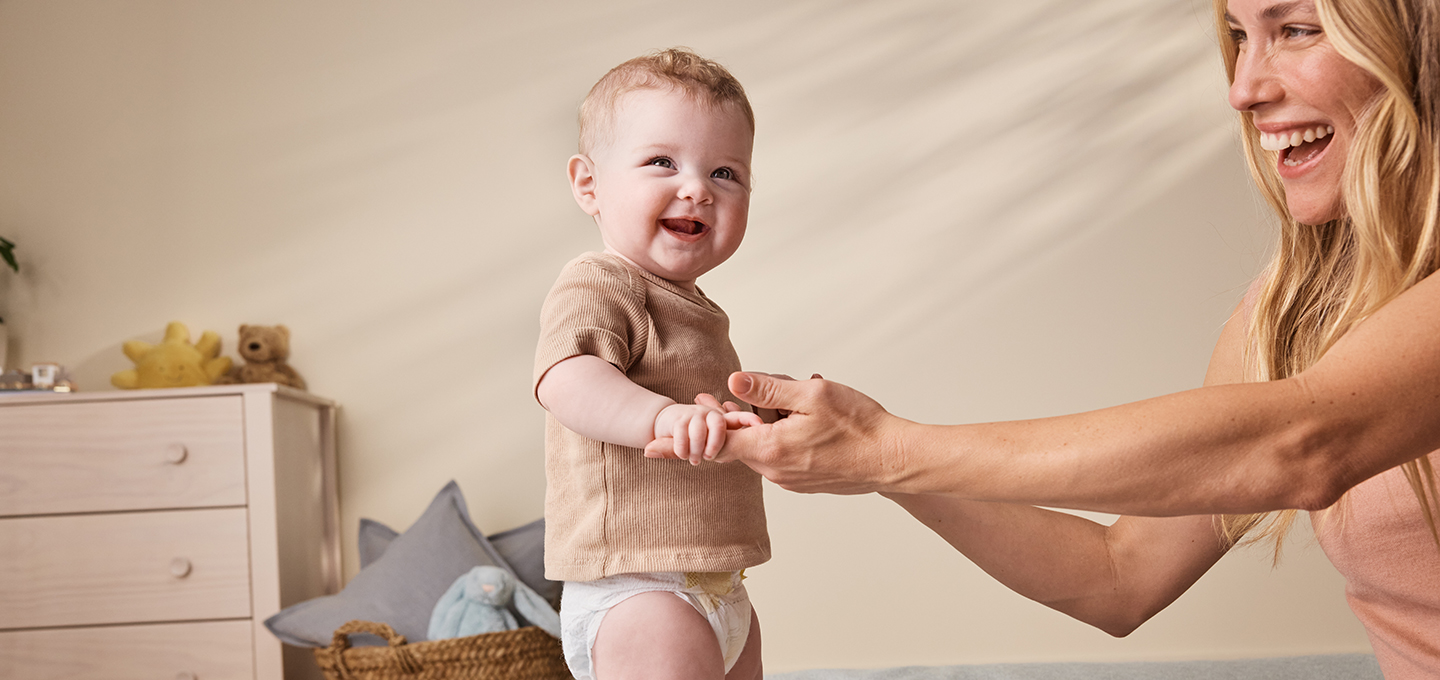 Smiling baby in a diaper stands on a bed holding an adult’s hands in a calm, cozy bedroom, showing gentle parenting support.]