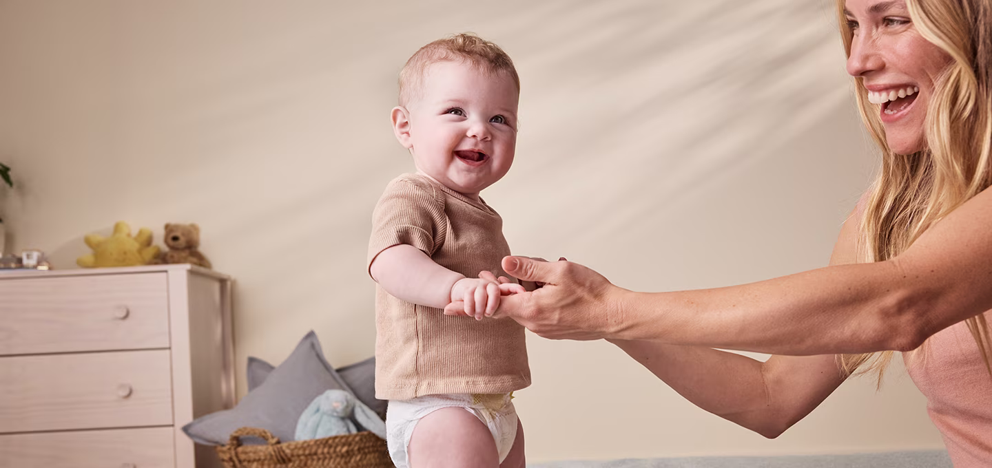 Smiling baby in a diaper stands on a bed holding an adult’s hands in a calm, cozy bedroom, showing gentle parenting support.]