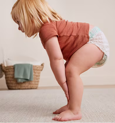 A toddler in a red shirt bending over to the side with her knees bent and hands on her legs, showing off the side of a Swaddlers 360 