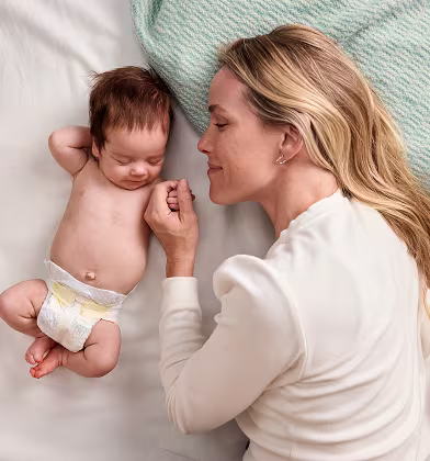 Baby laying peacefully on their back wearing Pampers Swaddlers and mom laying next to them holding their hand and smiling Baby laying peacefully on their back wearing Pampers Swaddlers and mom laying next to them holding their hand and smiling