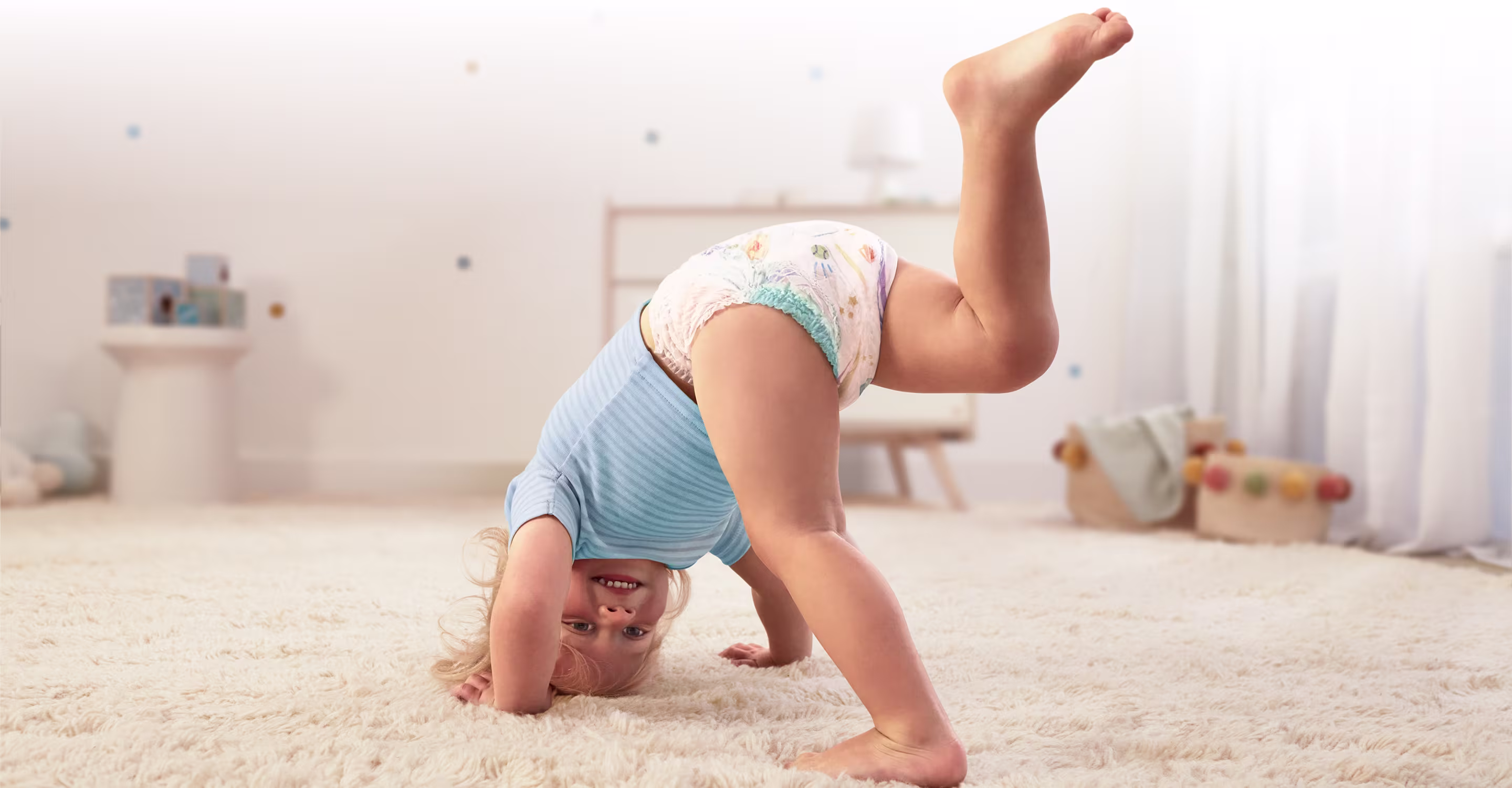 Ein glücklicher Kleinkind in Pampers Baby-Dry Pants macht einen Handstand auf einem flauschigen Teppich und lächelt in die Kamera.