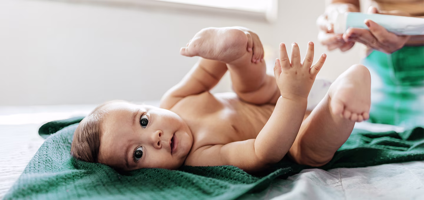 Baby lying on a green blanket during a diaper change as a caregiver prepares supplies, illustrating gentle home care.